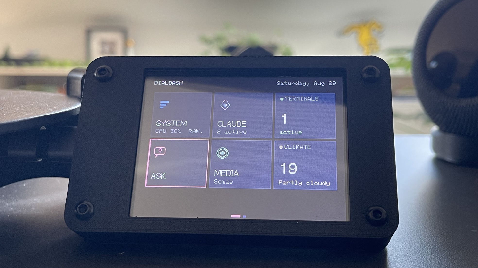 The DIALDASH home grid on the device, Saturday Aug 29: tiles for SYSTEM, CLAUDE with 2 active, TERMINALS with 1 active, ASK selected in pink, MEDIA playing Somae, and CLIMATE at 19 degrees partly cloudy.
